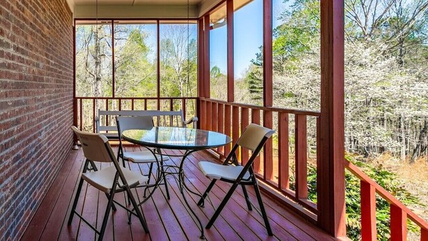 A covered outdoor area with a glass table and chairs, a brick wall, and a view of trees.