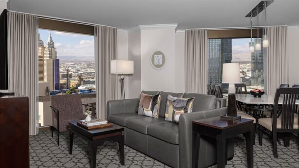 A modern hotel room with a gray sofa, a dining table, and a view of the cityscape.