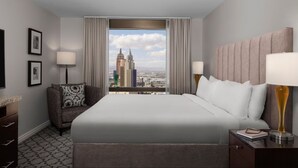 A hotel room with a large bed, a view of the cityscape, a sofa, and a nightstand with a lamp.