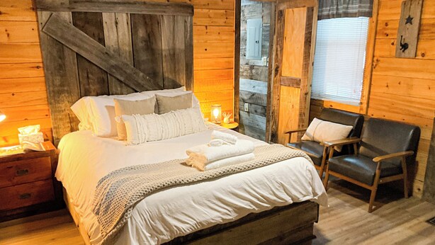 A wooden cabin bedroom with a large bed, bedside tables, a chair, and a window with blinds.