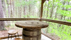 A rustic wooden table with a barrel base, two stools, and a metal bar stool on a wooden deck with a metal railing, overlooking a forested area.
