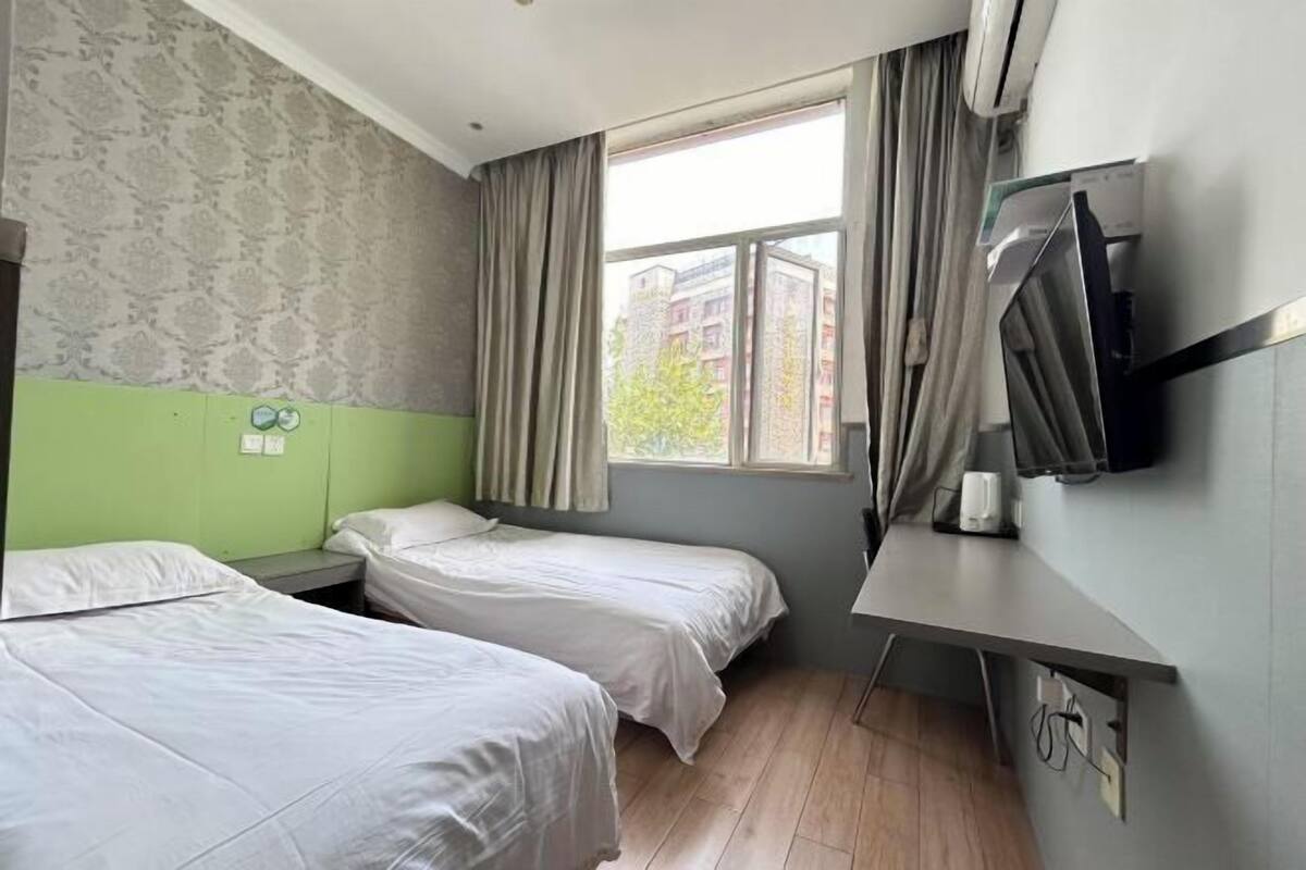 Habitación de hotel con dos camas individuales, un escritorio con silla, televisor montado en la pared y ventana con cortinas.