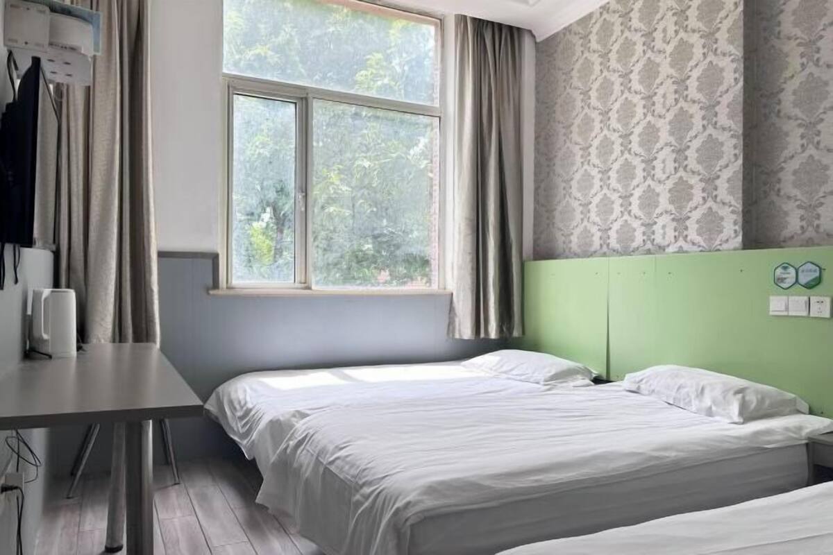 Habitación con dos camas, una ventana con cortinas, un cabecero verde y un papel tapiz estampado en la pared.