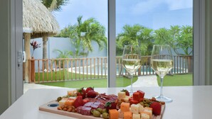 Un tavolo con un tagliere di salumi e due bicchieri di vino bianco, con vista su una piscina e un giardino tropicale.