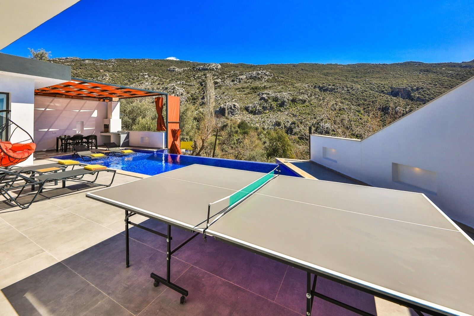 A rooftop pool area with a ping pong table, outdoor seating, and a view of a mountainous landscape.