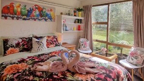 A hotel room with a floral bedspread, a painting of colorful birds on the wall, a bookshelf, and a view of greenery through the window.