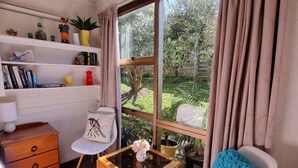 A cozy room with a wooden dresser, a white chair, a small table with a vase, and a large window with curtains.