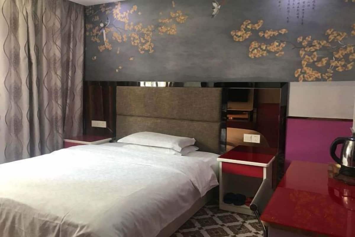Une chambre d’hôtel avec un lit, une table de chevet, une table rouge, un mur orné d’un motif floral et une étagère remplie de livres.