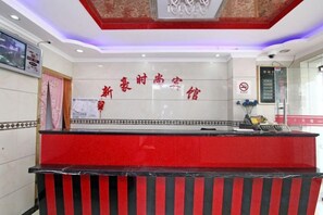 Uma área de recepção de hotel com um balcão vermelho, uma placa com caracteres chineses fixada na parede e uma câmera de segurança acima do balcão.