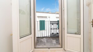 Une porte ouverte donnant sur un balcon d’où l’on peut admirer un bâtiment blanc aux volets verts.