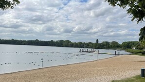 A sandy beach with a calm lake, a dock with boats, and a grassy area with trees. - Vacation apartment directly on the lake (Kalkar)