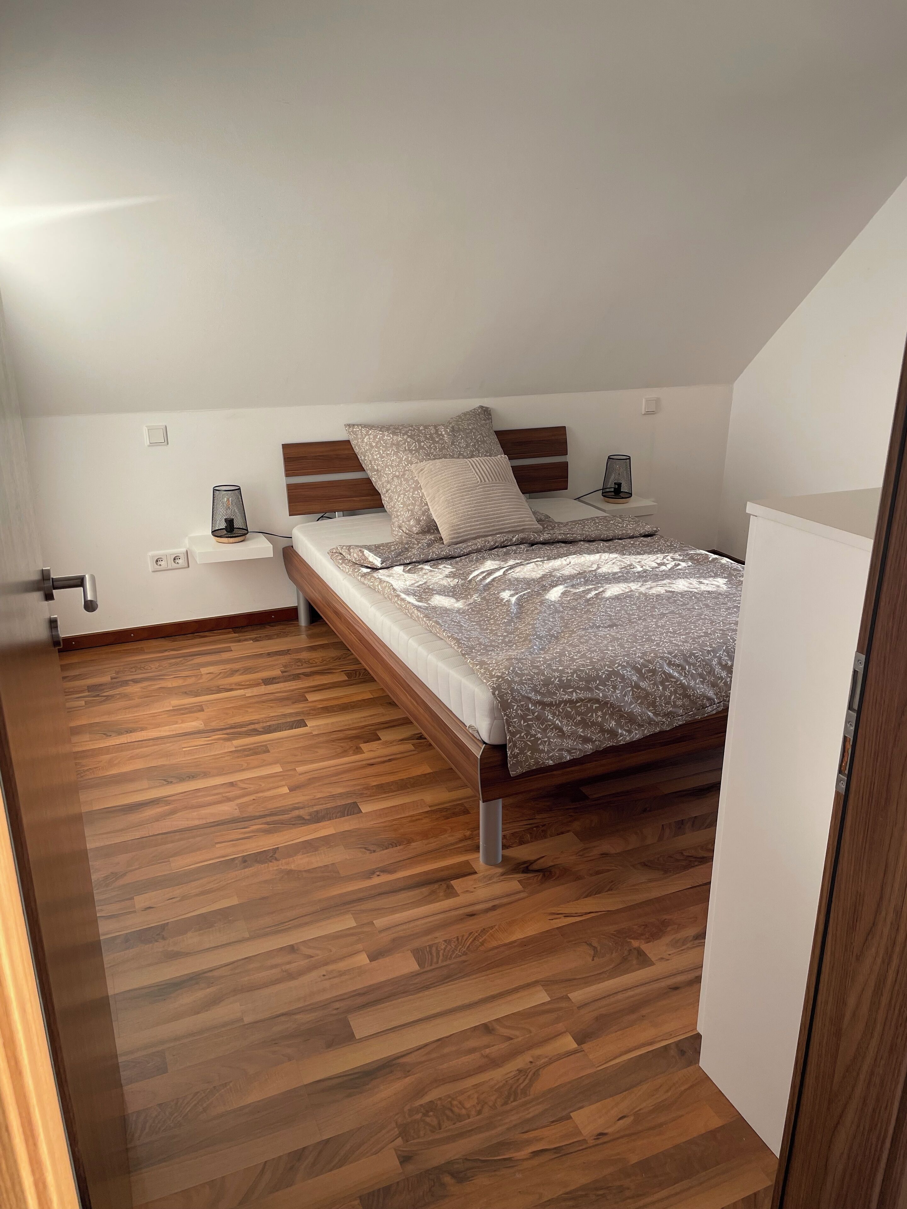 A bedroom with a wooden bed, a nightstand, a wall-mounted lamp, and a wooden floor.
