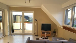 A modern living room with a sofa, a TV on a wooden stand, a dining table, and a large sliding glass door leading to an outdoor area. - Vacation apartment directly on the lake (Kalkar)