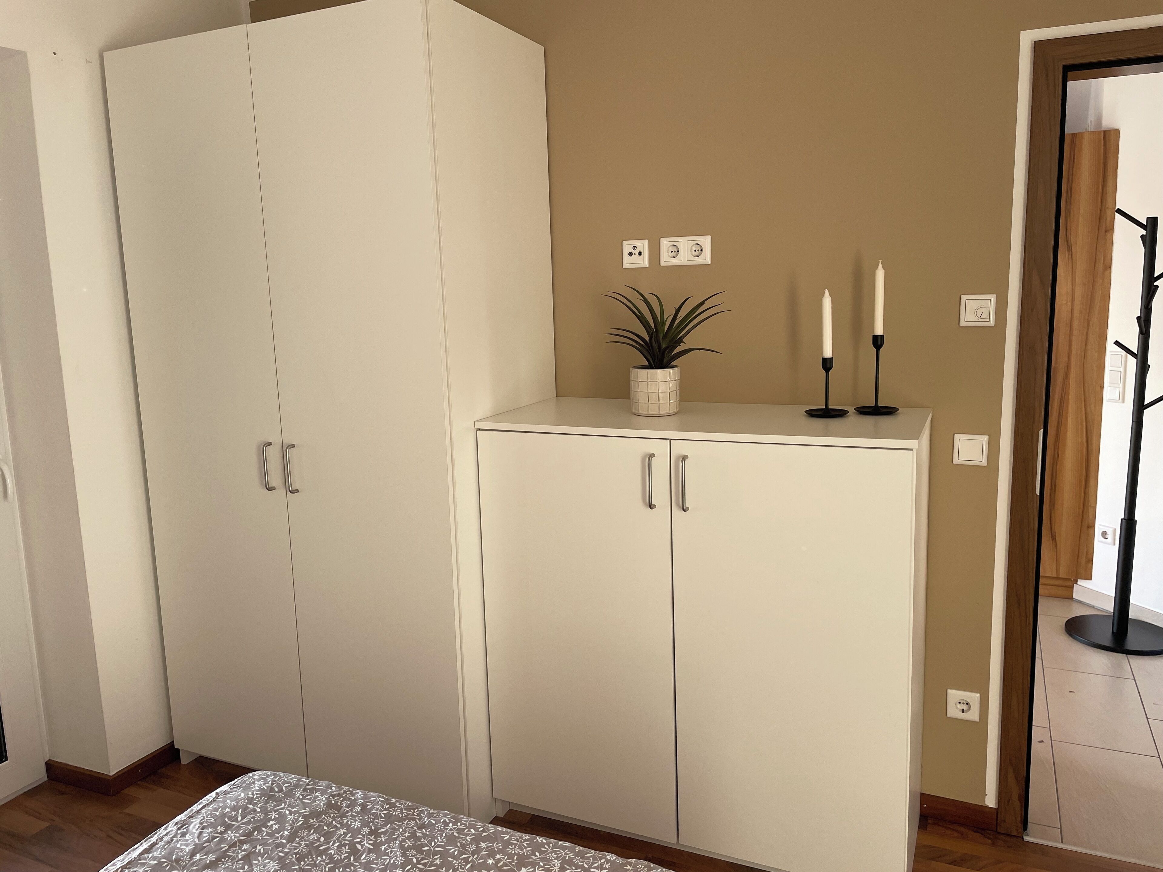Een kamer met een grote witte garderobe, een klein wit kastje met twee deuren, een potplant, twee kaarsen en een kapstok.