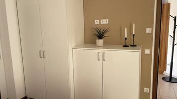 Een kamer met een grote witte garderobe, een klein wit kastje met twee deuren, een potplant, twee kaarsen en een kapstok.