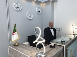 A hotel reception area with a modern design, featuring a wooden reception desk, a decorative lamp, and multiple clocks on the wall.