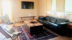 Un salon avec un canapé en cuir noir, une table basse en bois, un fauteuil blanc à poils longs, un téléviseur à écran plat fixé au mur et un tapis à motifs.