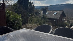 Eine Terrasse mit einem Glastisch und Stühlen, die auf eine ländliche Landschaft mit Bäumen und einem Haus blickt.
