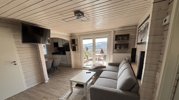 Ein gemütliches Wohnzimmer mit grauem Sofa, einem kleinen weißen Tisch und einem an der Wand befestigten Fernseher.