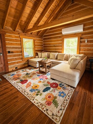 L’intérieur d’une cabane en rondins, avec un canapé d’angle, un tapis à motifs floraux et des poutres en bois.