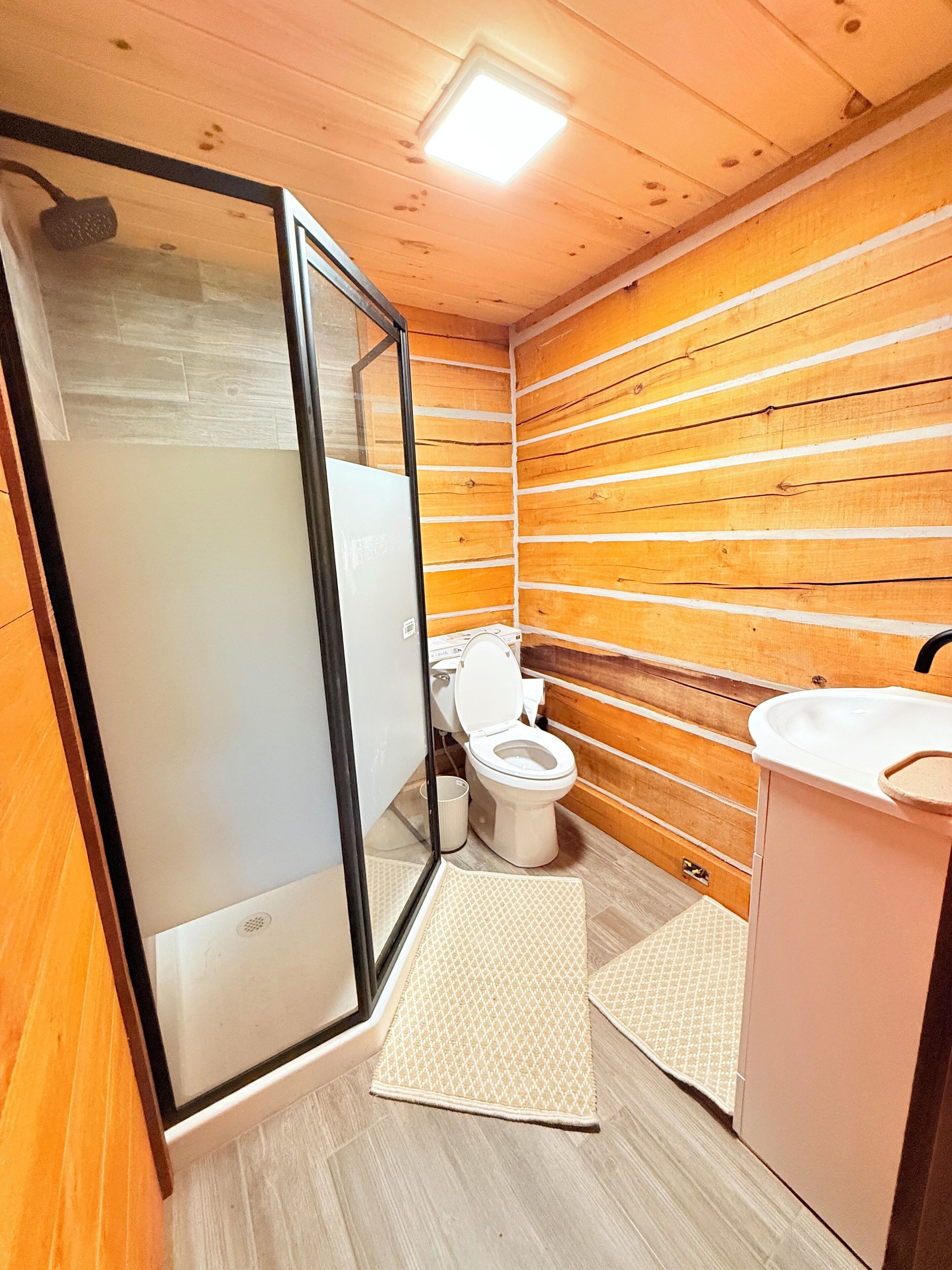 Une salle de bain avec des murs en bois, des toilettes, un lavabo et une douche dotée d’une porte en verre.