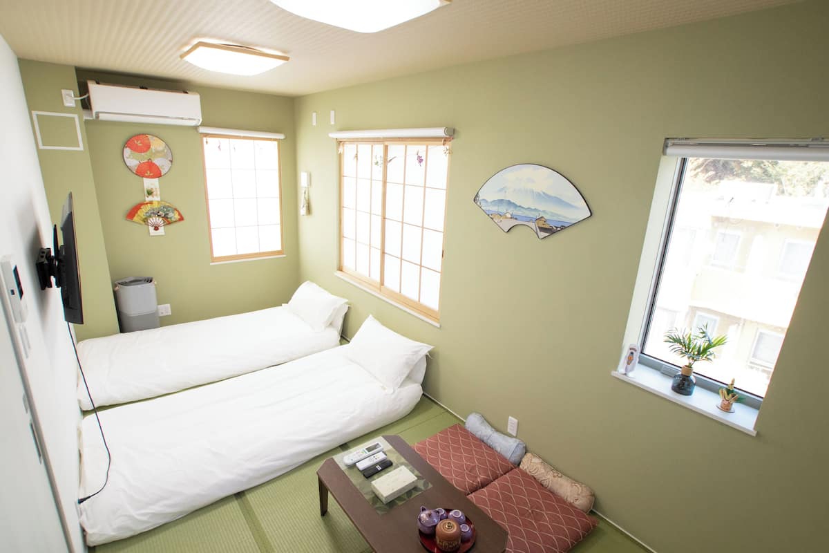 A room with two beds, a small table with a teapot, a window with a plant, and a wall clock.