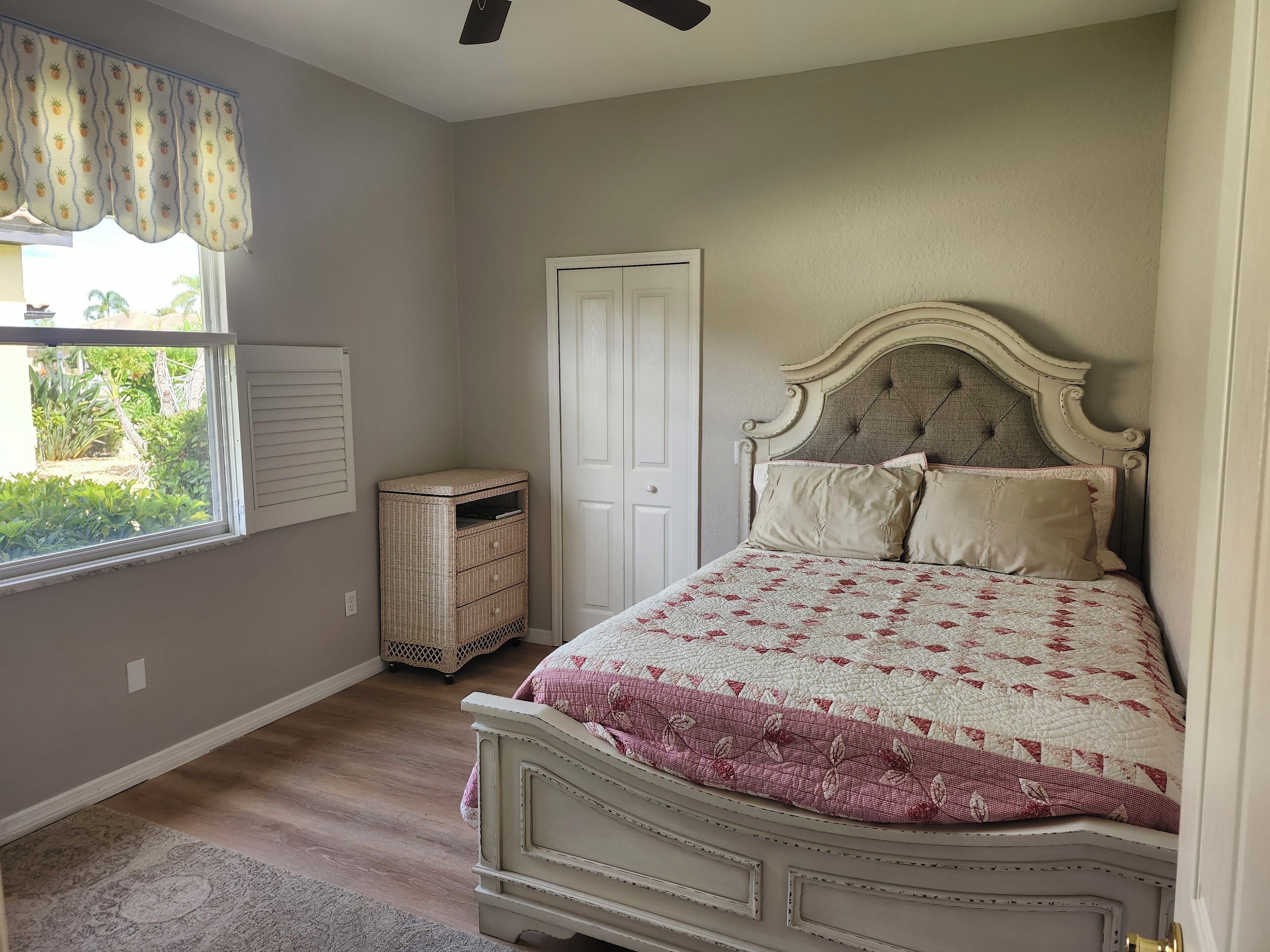 A bedroom with a bed, a window with a curtain, a door, and a small wicker chest of drawers.