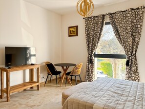 A room with a bed, a TV on a wooden table, two chairs, a patterned curtain, and a picture on the wall.
