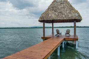 Een houten steiger die zich uitstrekt over het water, met een rieten dakgebouw aan het einde, twee stoelen en uitzicht op land in de verte.