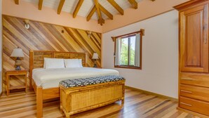 Un dormitorio con cama de madera, mesitas de noche, un banco de mimbre, una ventana con cortinas y un armario.