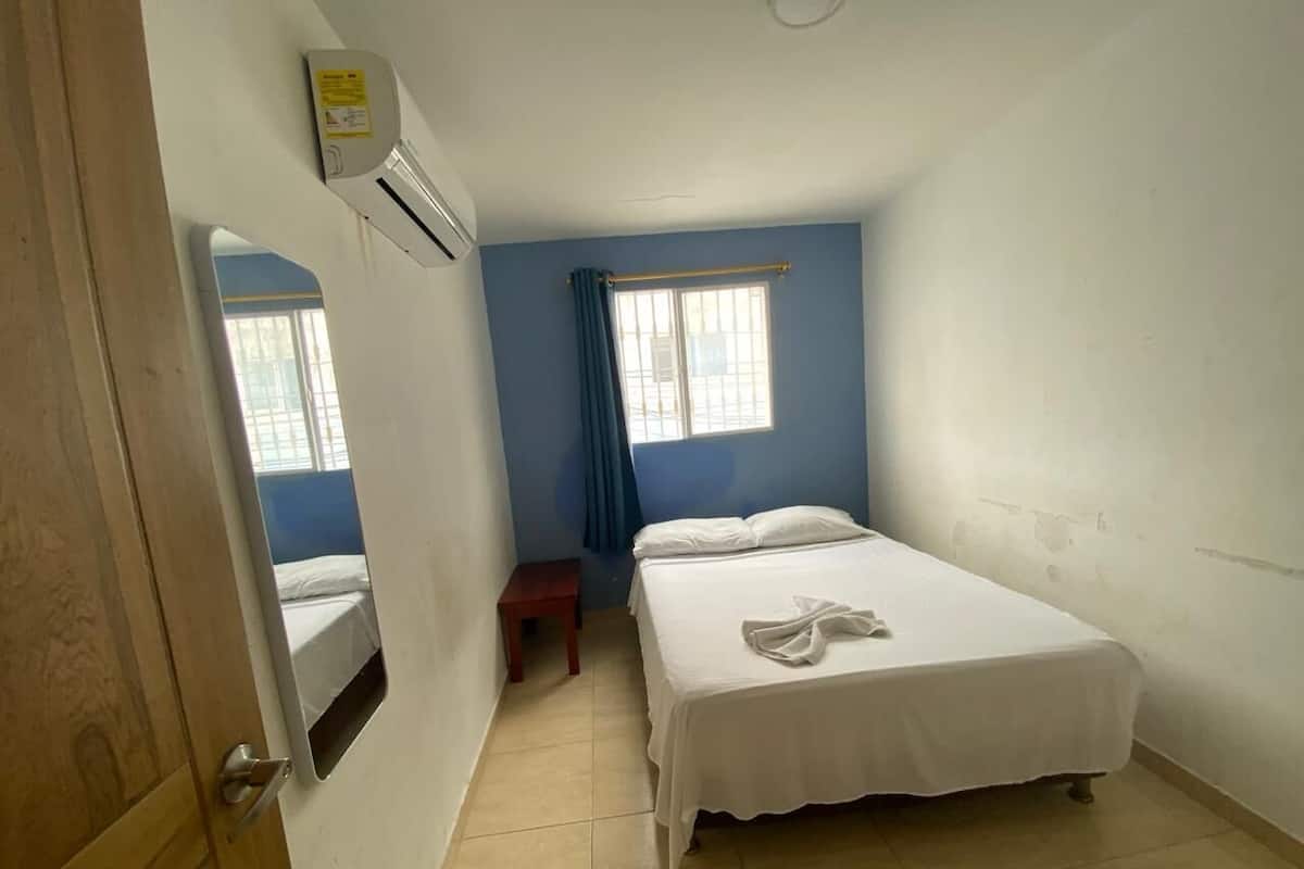 A small, clean room with a bed, a window with curtains, and an air conditioning unit.