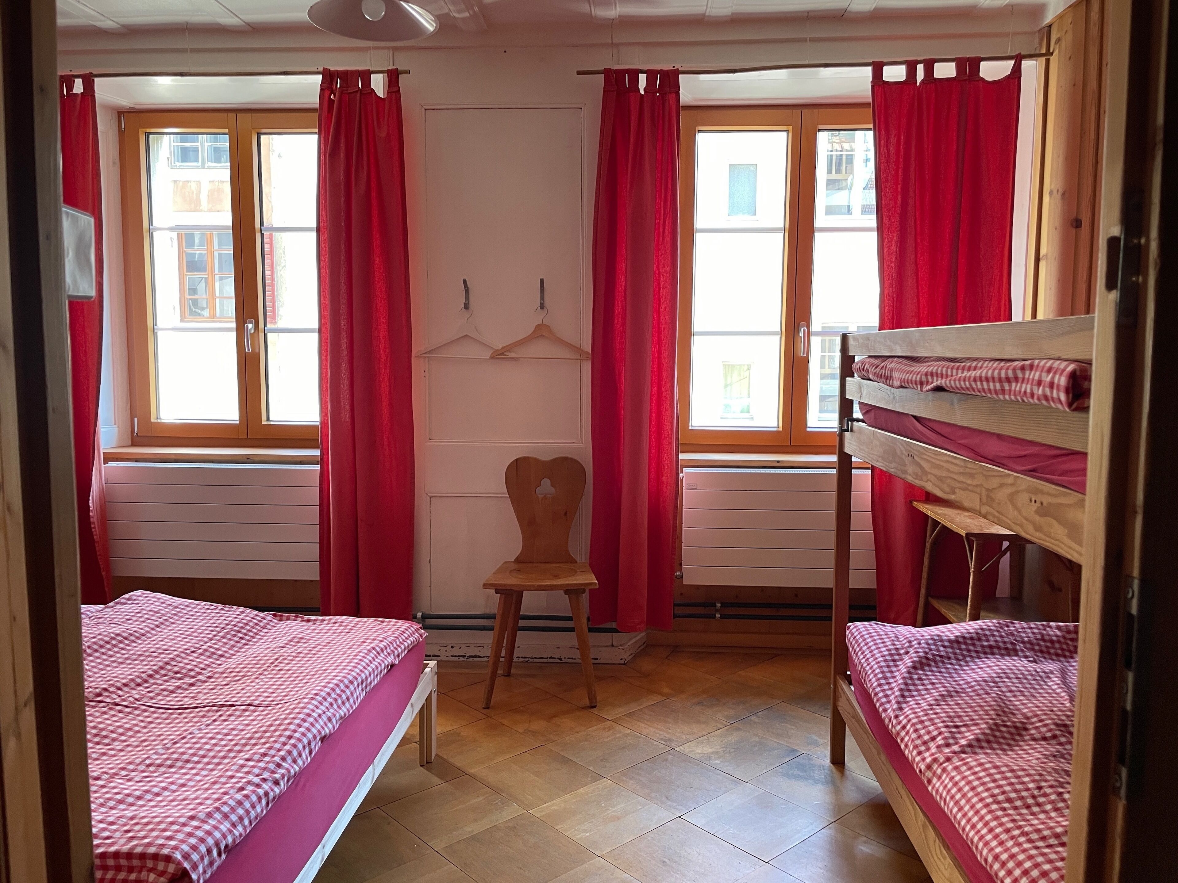 Een kamer met twee bedden, een stoel en een raam met rode gordijnen.