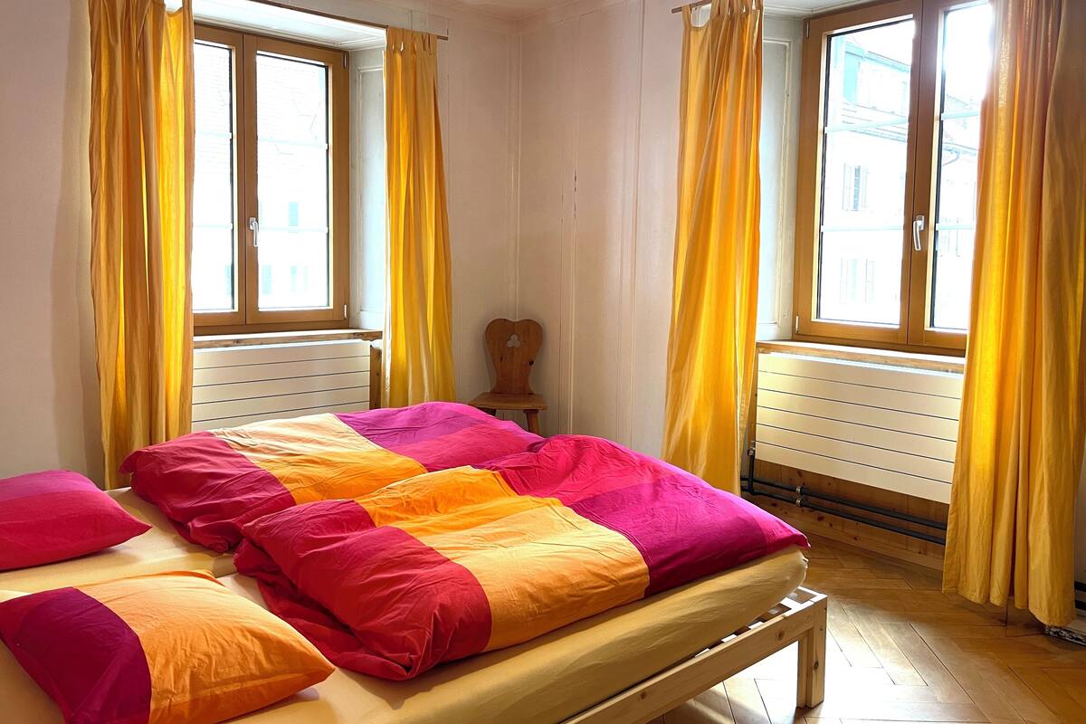 A room with a bed, two windows with yellow curtains, a wooden chair, and a radiator.