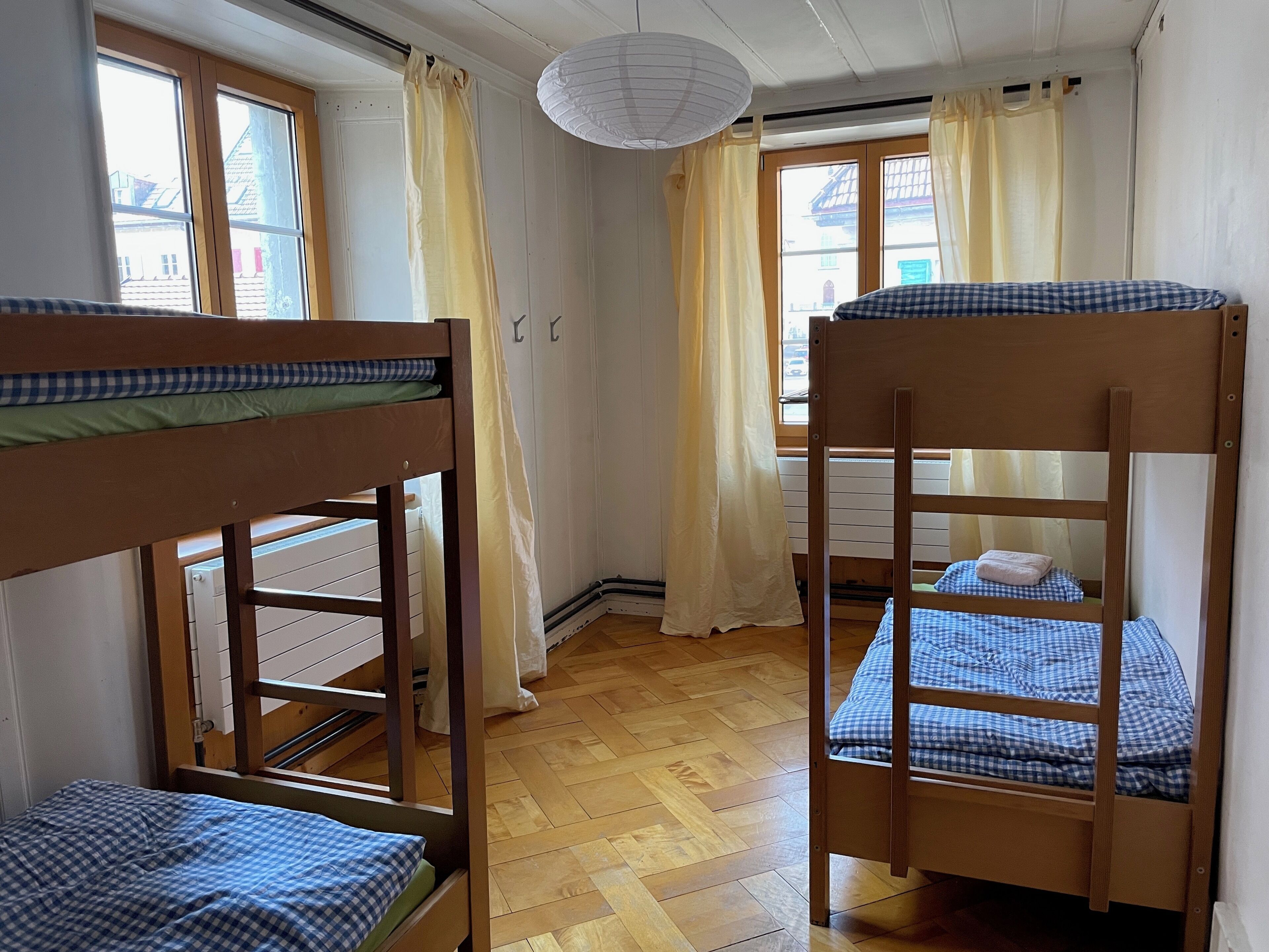 A room with bunk beds, wooden flooring, and large windows with curtains.