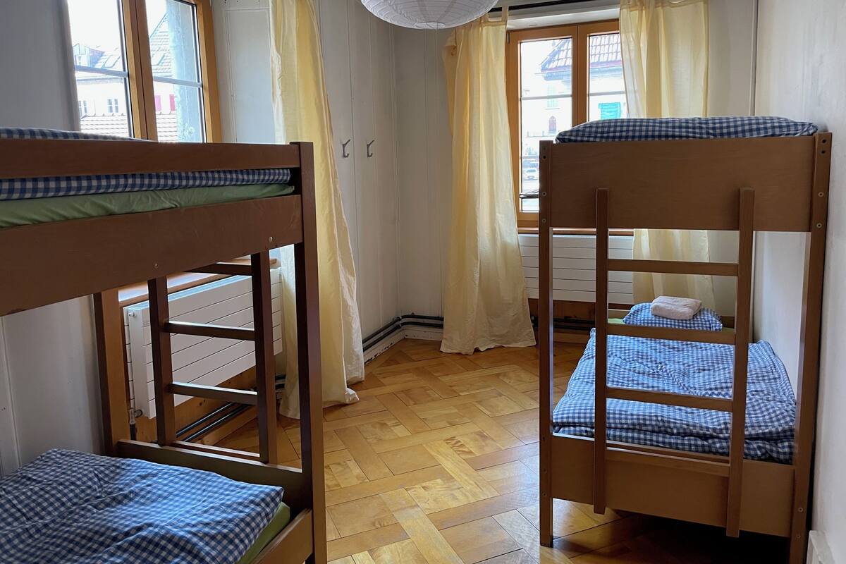 A room with bunk beds, wooden flooring, and large windows with curtains.