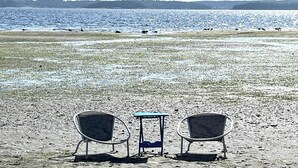 Two metal chairs and a small table on a sandy beach with a view of the ocean and distant trees. - Oceanfront, beach access Cozy bed & breakfast loaded  amenities , Sunny Saltair (Ladysmith)