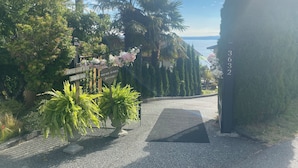 A pathway leading to a house with a view of the sea, surrounded by lush greenery and flowering plants. - Oceanfront, beach access Cozy bed & breakfast loaded  amenities , Sunny Saltair (Ladysmith)