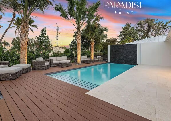 Un espace piscine extérieur moderne, doté d’une terrasse en bois, de chaises longues et de palmiers.