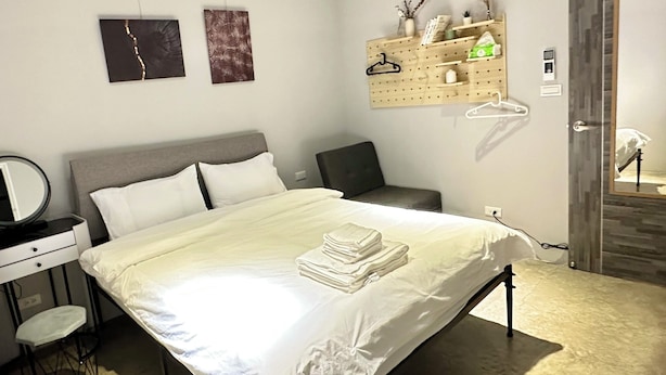 Een keurig opgemaakt slaapkamer met een bed, nachtkastje, stoel en een wandplank met diverse spullen.