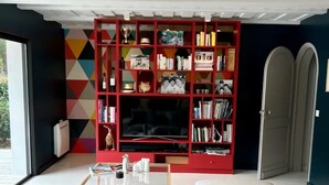 Un salon moderne avec un grand canapé gris, une bibliothèque rouge remplie de livres et d’objets décoratifs, un téléviseur à écran plat et une grande fenêtre offrant une vue sur l’extérieur.