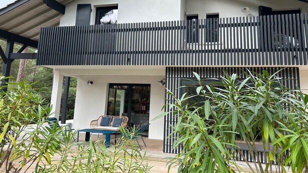 Une maison moderne à deux étages, dotée d’un balcon, d’un espace extérieur aménagé pour s’asseoir et de plantes en pot.