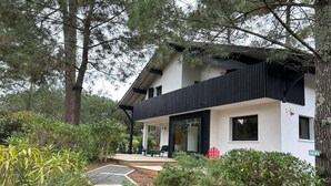 Une maison moderne à deux étages, dotée d’un porche abrité, entourée d’une végétation luxuriante et de grands arbres.
