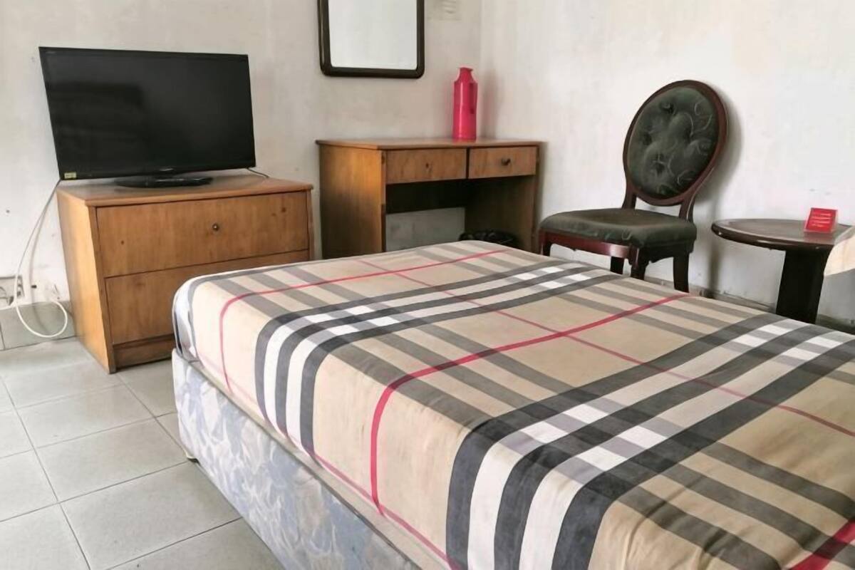 Quarto com cama, cómoda de madeira, televisão, secretária, cadeira e uma pequena mesa.