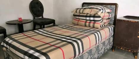A single bed with a patterned bedspread, a bedside table, a chair, and a small table with a red object on it.