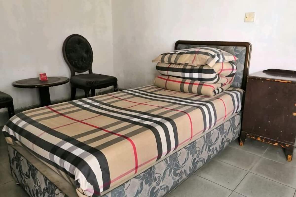 Uma cama de solteiro com colcha xadrez, mesa de cabeceira, uma cadeira e uma pequena mesa com um objeto vermelho.
