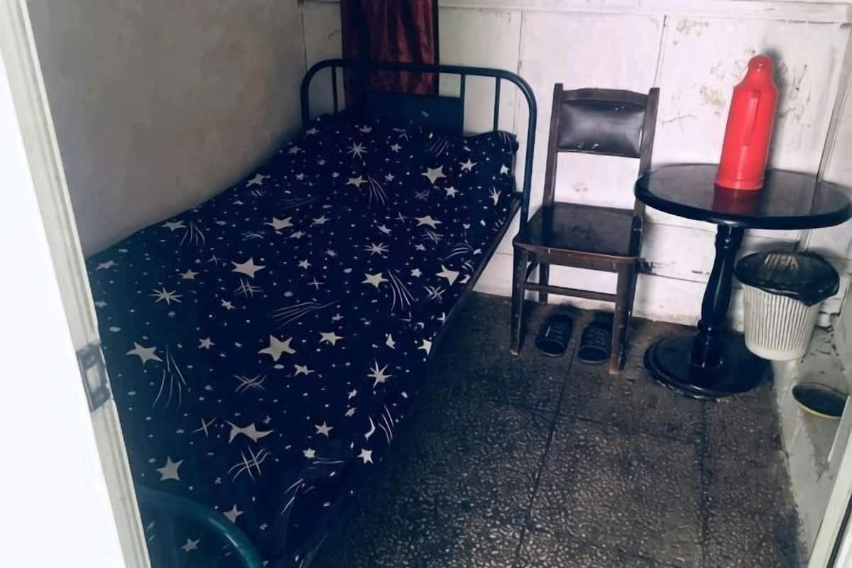 A small, single-bed room with a star-patterned bedspread, a chair, a small round table, and a red bottle.