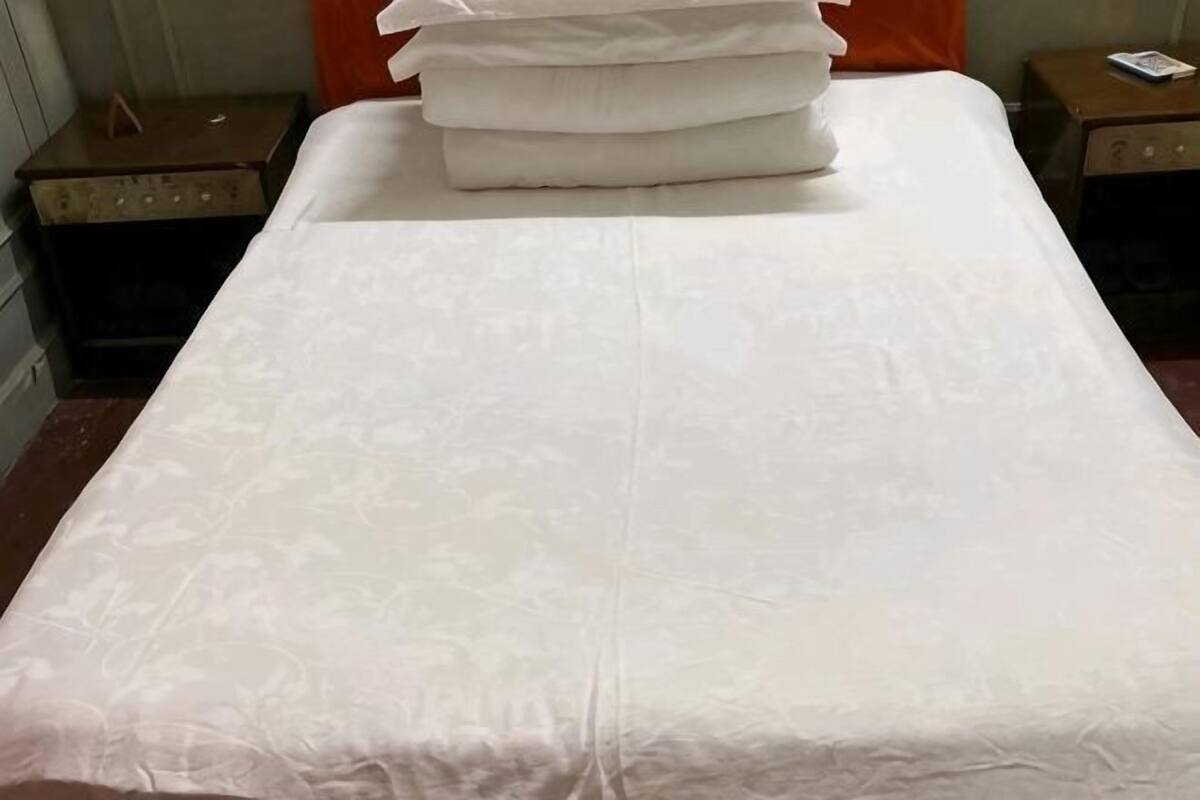 Uma cama bem arrumada com um cobre-leito branco e almofadas, uma mala à esquerda e uma mesa de cabeceira com um abajur à direita.