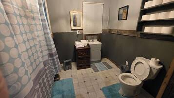 A bathroom with a toilet, sink, and a mirror. There are towels stacked on the counter, and a trash can is visible.