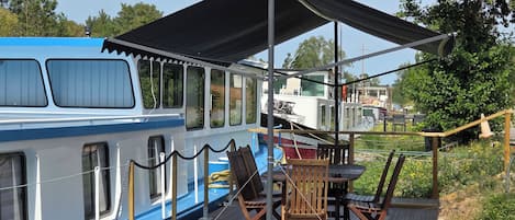 En træterrasse med et bord og stole, med udsigt over en flod med både.
