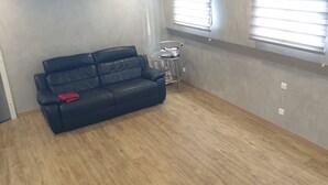 Een kamer met een donkerblauwe leren bank, een klein metalen tafeltje en een raam met zonwering.